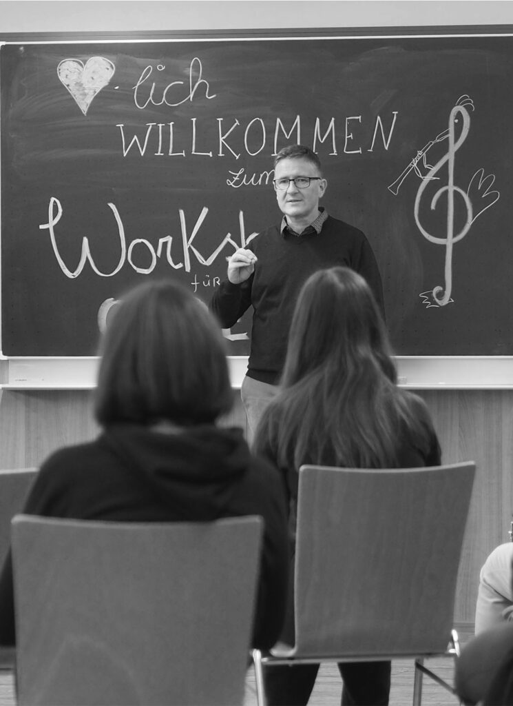 Fabio vor einer Tafel auf der Herzlich Willkommen zum Workshop steht, davor sitzen Teilnehmer